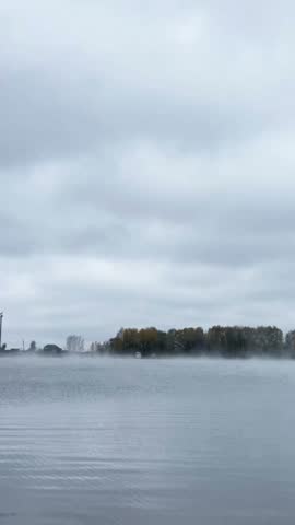 Calm autumn lake covered with low morning mist, distant trees and a dramatic overcast sky.