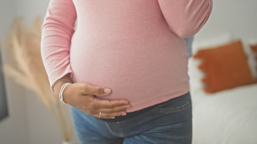 Pregnant woman hugged from behind by man with arms around her shoulders and belly in building; joyful anticipation.