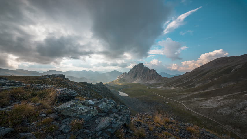 Fast-moving clouds cross the sky above a jagged mountain and broad alpine basin with rocky foreground and winding trail.