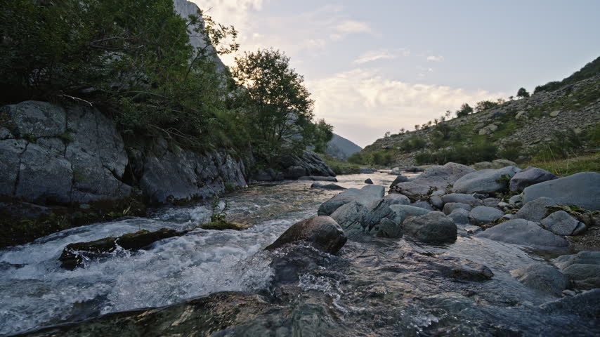 Shallow stream water ripples over smooth rocks in a narrow green gorge at dusk, framed by trees and distant hills.