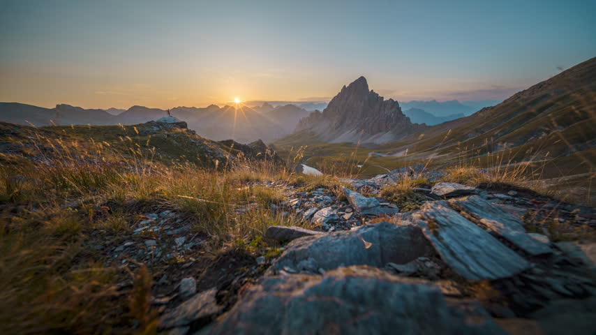 Dawn light grows behind a sharp central peak as grasses and rocks in the high valley foreground slowly brighten.