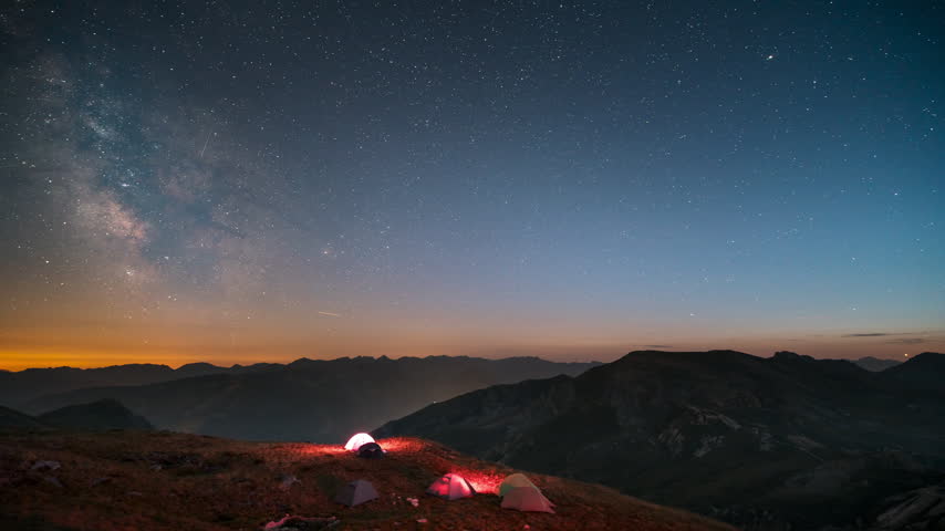 Backcountry tents sit on a dark ridge as the Milky Way core climbs above distant silhouettes in a night sky timelapse.