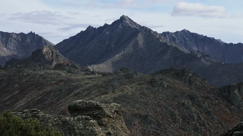 Jagged mountain peaks rise above scattered boulders and sparse vegetation as drifting clouds cast shifting shadows across the rugged, layered ridges under a partly cloudy sky.