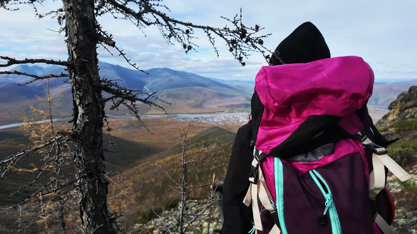 A hiker in a black beanie and pink backpack stands still on a rocky overlook, gazing at a valley with rolling hills, a river, and a distant town under a partly cloudy sky.