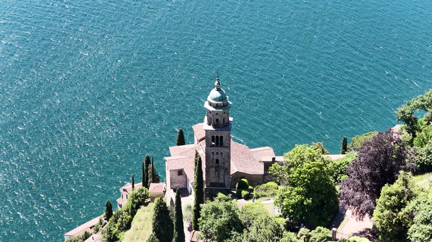 Santa Maria del Sasso church perches above deep blue Lake Lugano in Morcote, Switzerland, aerial drone orbit around bell tower and lakeside chapel.