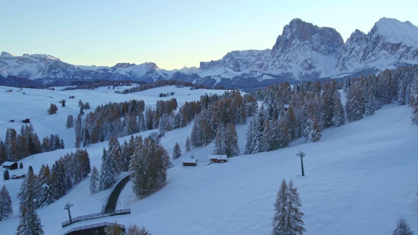 Peaceful snowy mountain vista. Alpine trees dusted with snow. Ski lift. Sunrise glow over majestic peaks. Drone footage.