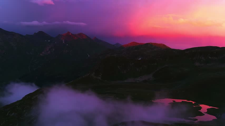 Dramatic aerial view of rocky mountain ridges under glowing pink and purple dusk clouds. Ethereal fog drifts through dark valleys over calm glacial lakes in scenic wilderness scene.