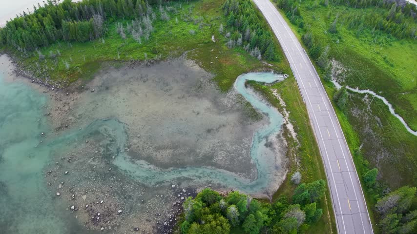 Top down drone footage shows a paved road curving beside shallow turquoise wetland water and green marsh vegetation.