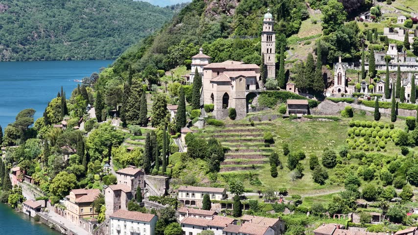 Santa Maria del Sasso church stands on terraced hillside above Lake Lugano in Morcote, Switzerland, aerial drone shot of bell tower and village.