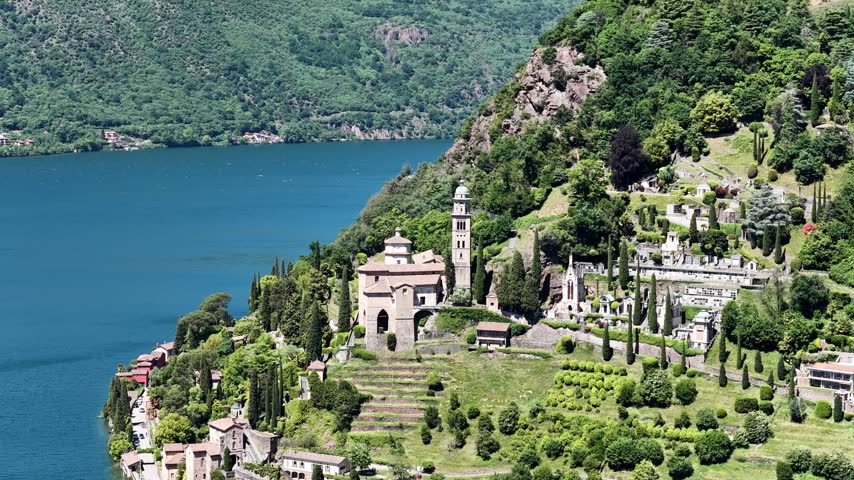 Santa Maria del Sasso church overlooks Lake Lugano in Morcote, Switzerland, aerial drone shot of bell tower, cemetery gardens and turquoise water.