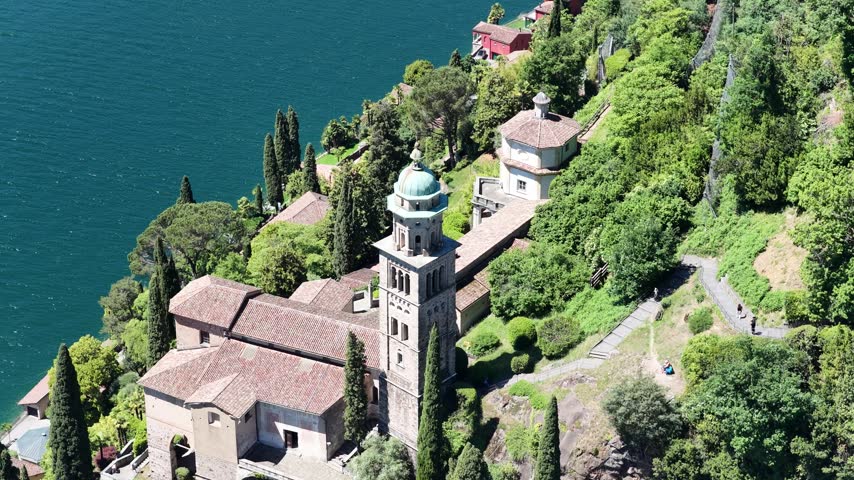 Santa Maria del Sasso church sits above Lake Lugano shoreline in Morcote, Switzerland, aerial drone pulls back from bell tower and gardens.