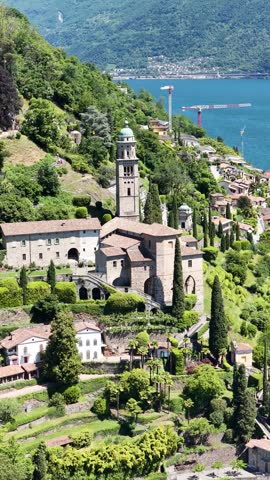 Santa Maria del Sasso church sits above Lake Lugano in Morcote, Switzerland, aerial drone orbit view, bell tower, gardens and Ticino hillside village.