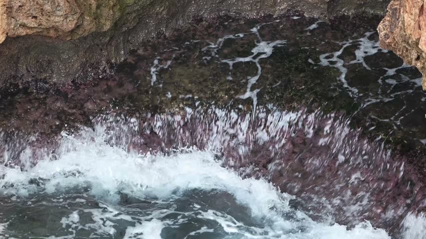 Waves roll in, hitting the rocky shore with force. Water splashes up creating foam and mist, showcasing nature’s raw power near the ocean coast.