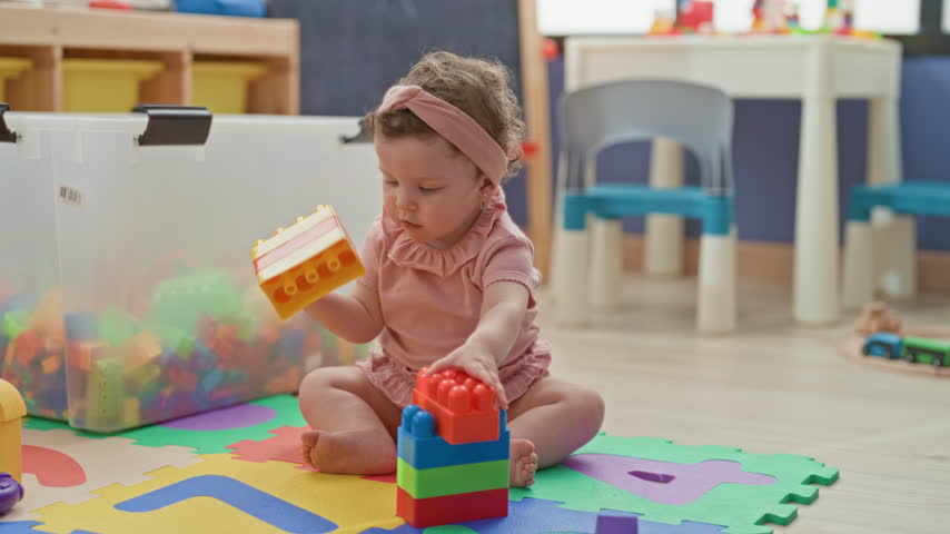 Toddler girl holding large plastic blocks, mouthing a red brick and sitting barefoot on a foam play mat in a daycare building; curiosity focus.