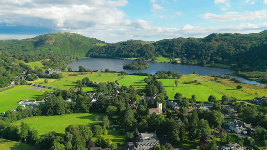 Lush green hills surround a calm lake under bright skies. Village nestled among trees and fields, peaceful and quiet. Soft clouds drift above. Grasmere is a village in the Lake District