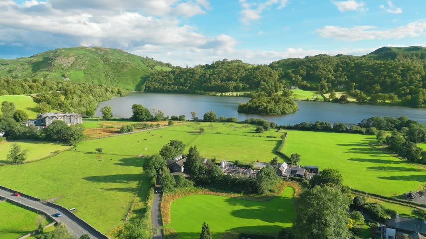 Lush green fields surround calm lake under bright blue sky. Pasture grazes cattle, ancient stone building stands nearby. Grasmere is a village in the Lake District