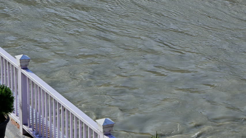 A potted shrub and diagonal white terrace railing overlook the textured surface of the Ganga River in Rishikesh.