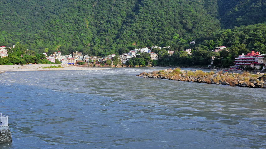 A broad stretch of the Ganga River passes a rocky shelf below wooded hills and buildings in Rishikesh.