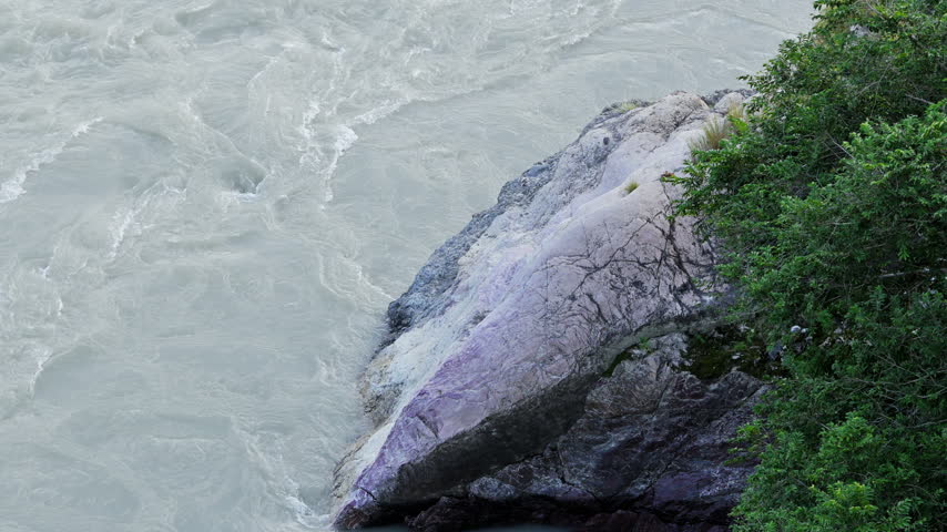 The Ganga River flows beside a large smooth boulder and overhanging foliage in Rishikesh.