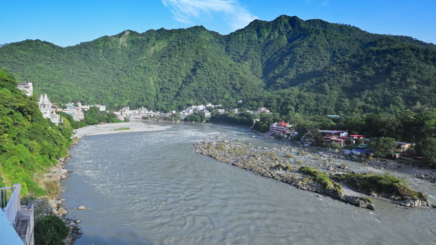 The Ganga River curves below a green hillside and riverside buildings in Rishikesh.
