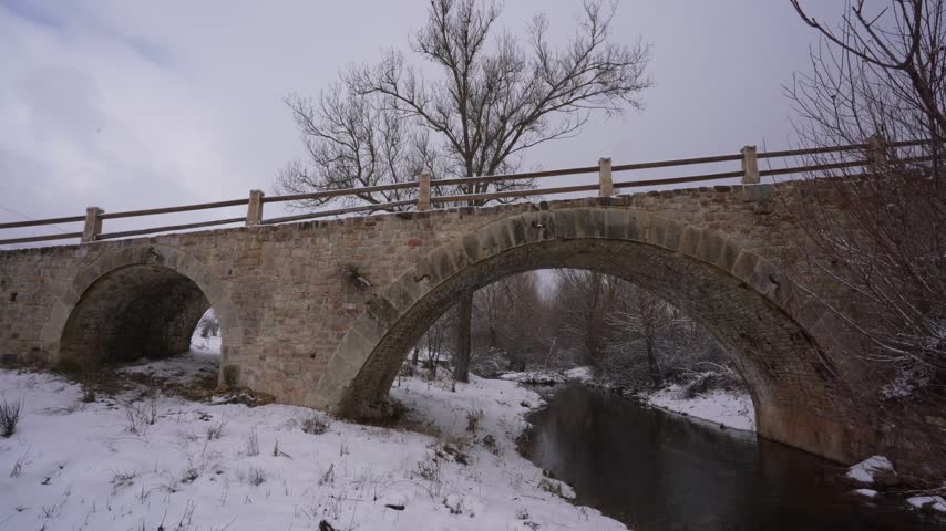 Ancient arched stone bridge crossing small calm river during winter day with snow covered landscape and bare trees backdrop.