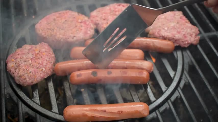 Cooking takes place outdoors with burgers and hot dogs on a grill. Smoke rises as the meat cooks, creating an enjoyable barbecue atmosphere with family.
