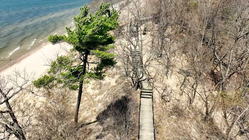 Aerial drone footage showing wooden stairs descending down sandy dune landscape towards blue lake shoreline during sunny day.