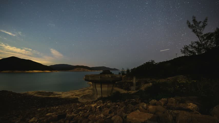 Dynamic time-lapse footage captures star trails moving across night sky over tranquil lake, hills, and a water intake structure.