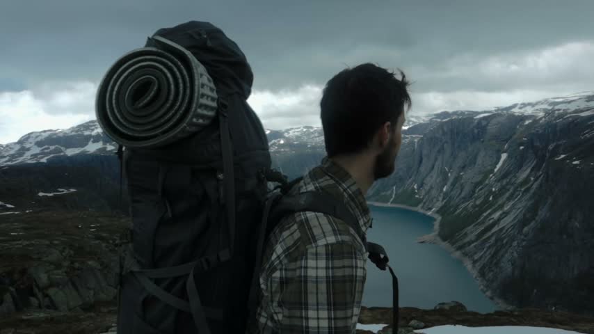 Young man backpacker standing on edge of cliff gazing at stunning mountain landscape and alpine lake scenic vista travel.
