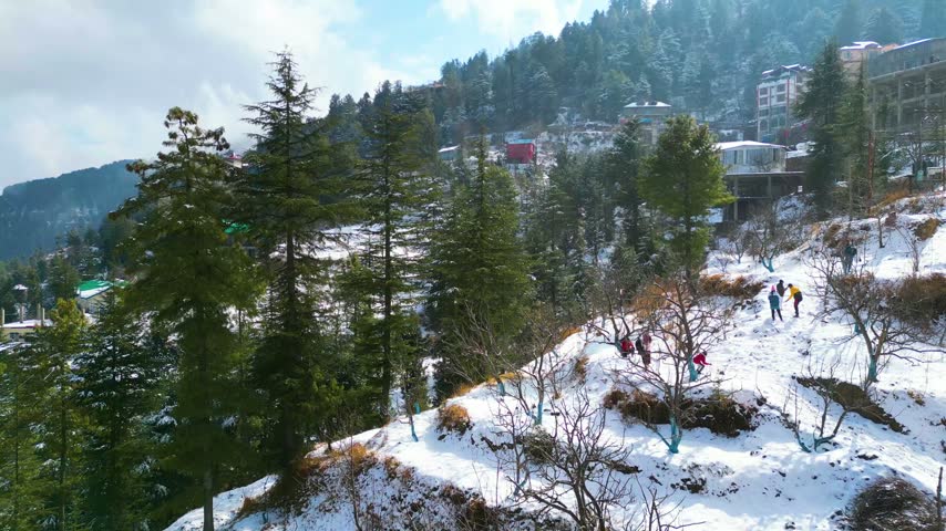Aerial view of snow covered hillside in mountain region with group of people walking and playing during sunny winter day in popular resort destination.