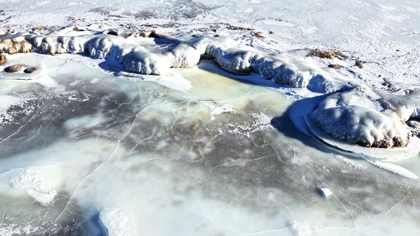Aerial view of a frozen lake with cracked ice patterns and icy rocks along the shore in winter