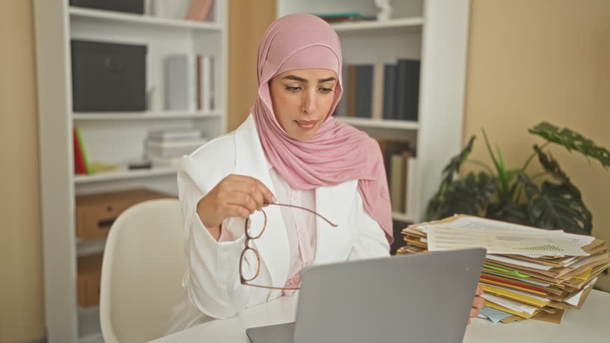 Woman holding glasses over laptop amid stacked files at a desk in building; confidence productivity.