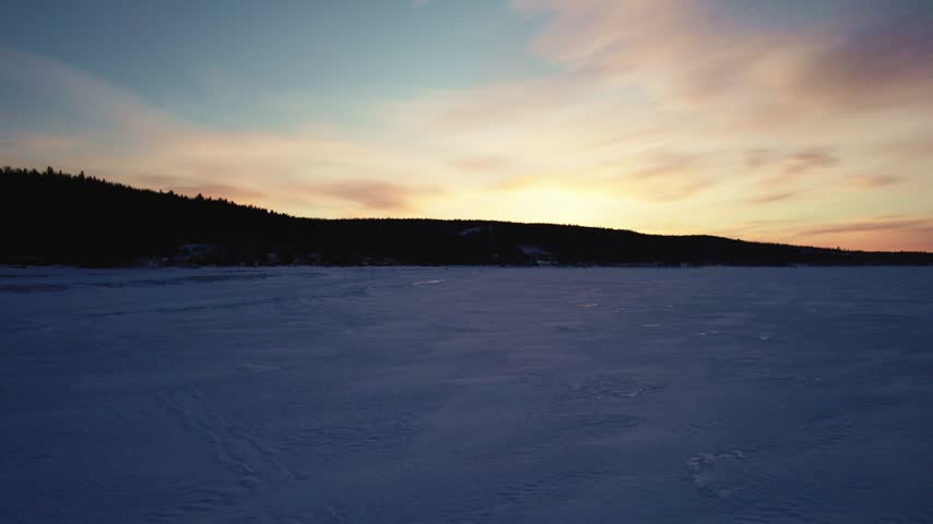 Aerial view of couple walking on ice covered lake at golden hour sunset with silhouettes against scenic Nordic landscape.