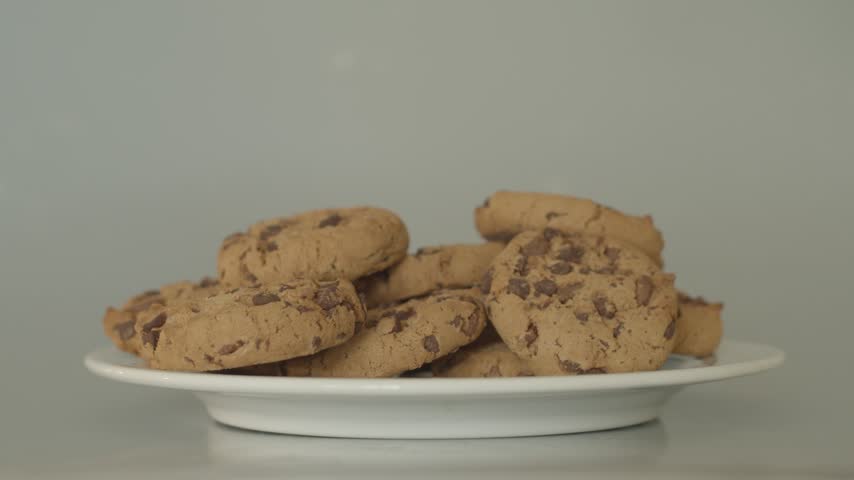 Hand adding delicious chocolate chip cookie to pile of baked snacks on white plate against neutral background. Close up food footage.