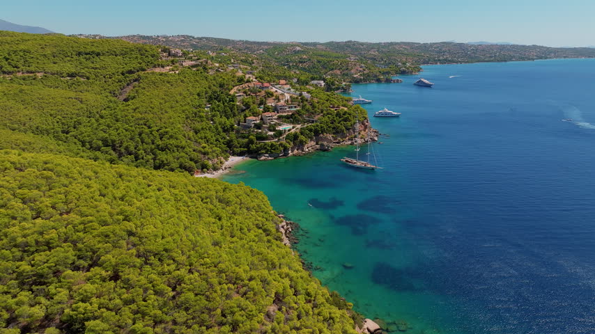 Aerial view of the pine-forested coastline of Kranidi, Greece, with Ormos Luxury Villas perched on the cliffs and yachts anchored in the turquoise Aegean below.