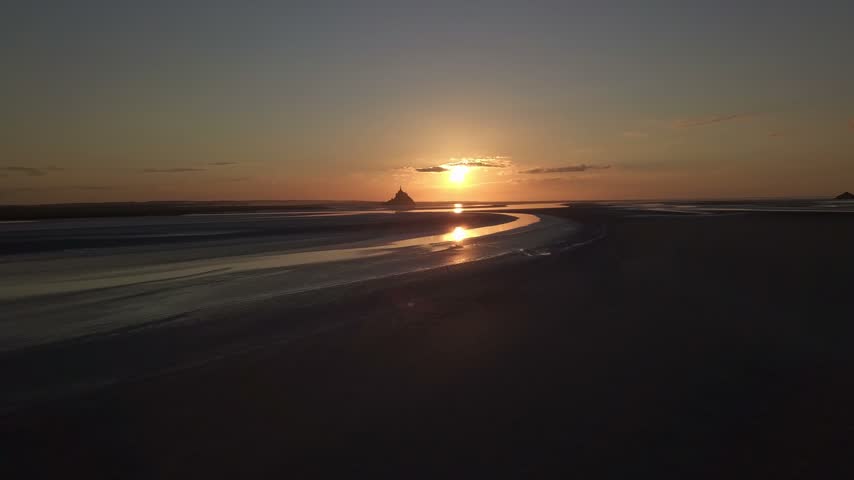 Breathtaking aerial view of Mont Saint Michel at sunset featuring calm tide and reflection of sun on wet sand in France.