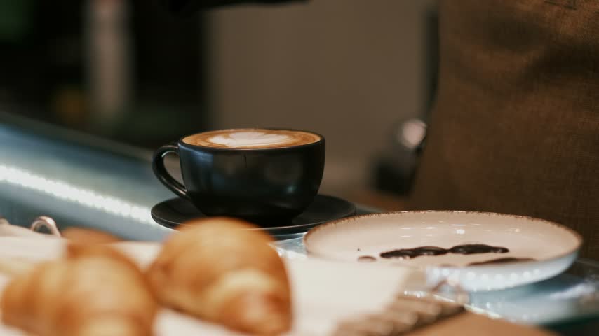 Gloved hand dips a sweet croissant into hot coffee making foam overflow on a cup in slow motion close up.