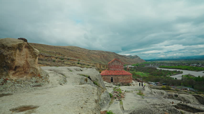 Visitors climb the worn rock path toward the red roofed basilica of Uplistsikhe above the wide green river valley.