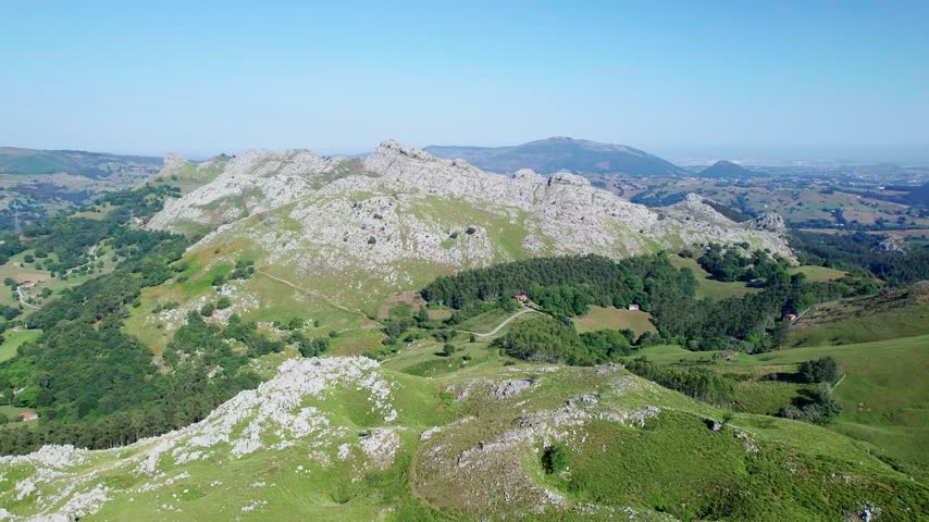 Cinematic aerial landscape showing rocky mountain ridges verdant slopes and winding paths under clear blue summer sky.