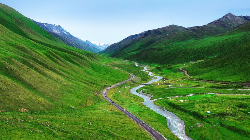 Aerial view of a winding river and highway crossing a vivid green mountain valley beneath rugged snowcapped peaks in Xinjiang.