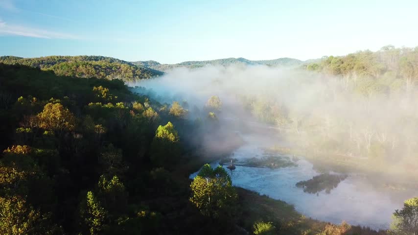 Aerial view over foggy river winding through forested Appalachian mountainside during calm golden hour morning sunlight.