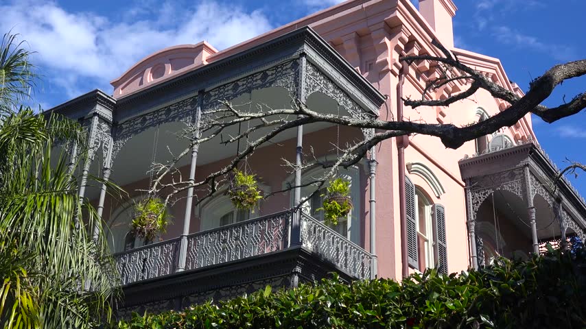 Historical pink mansion featuring intricate iron balcony and lush hanging plants set against clear blue sky in New Orleans.