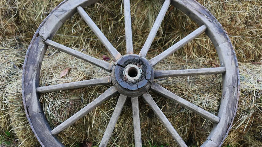 Old wooden wagon wheel leaning against hay bales on farm. Vintage rustic cart wheel with spokes. Rural countryside background, agriculture and traditional farming concept
