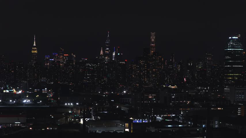 Aerial pan over the glowing Manhattan skyline illuminated against the night sky in New York City, revealing iconic skyscrapers and illuminated architectural towers across the urban landscape.