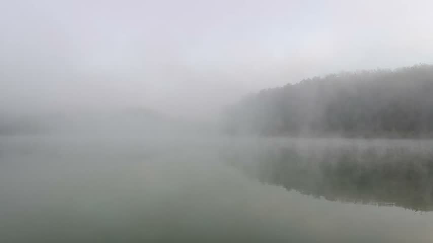 Thick morning fog rolling over a calm lake with forest reflection