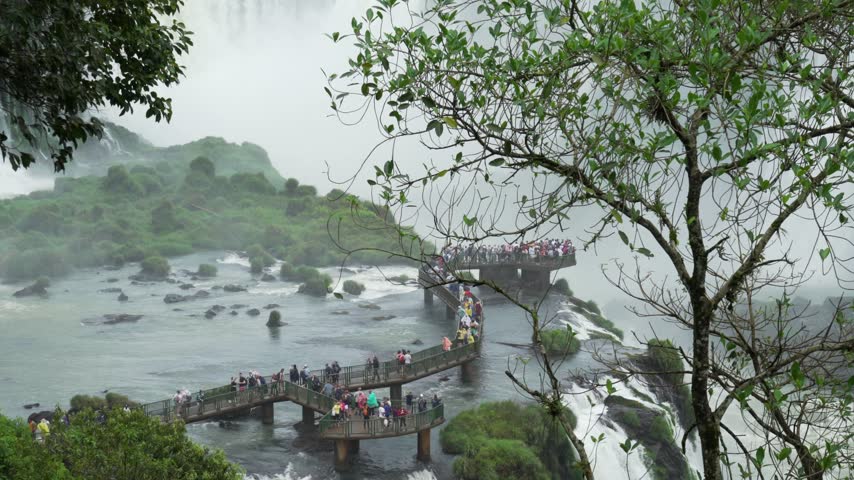 Crowd tourists crossing metal walkway over rushing water of Iguazu waterfalls surrounded by lush greenery in national park.