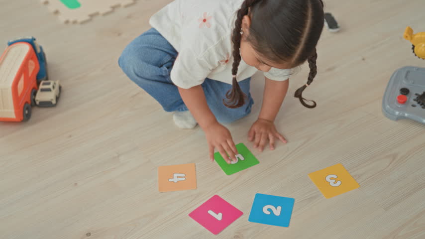Little hispanic girl sits on floor touching number cards and toy car with hand, legs crossed, playing in a building; curiosity and learning.