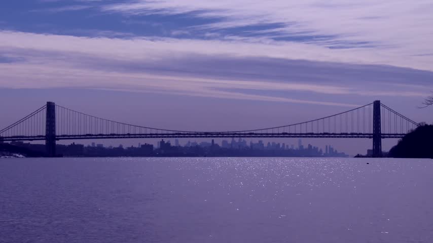 Iconic George Washington bridge suspended over calm Hudson river waters with hazy New York city skyline silhouette at sunset.