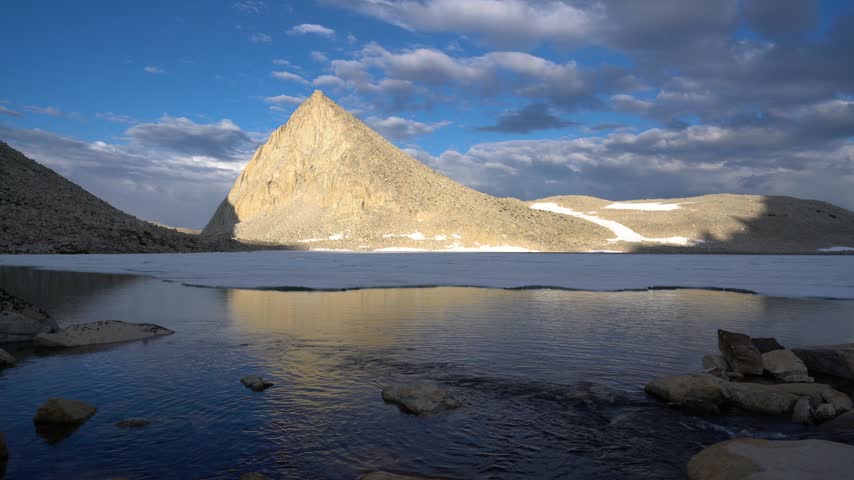 Stunning landscape shot features tranquil mountain lake with icy surface under dramatic clouds and jagged summit during golden hour light.