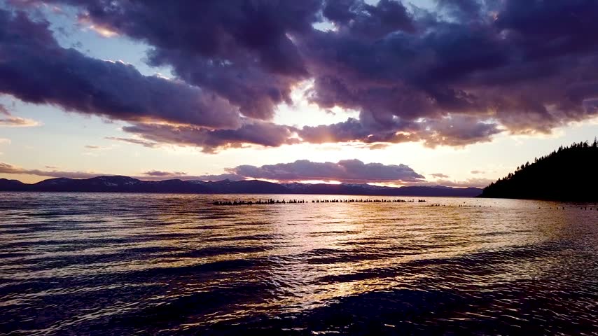 Slow motion shot of rippling lake water surface at sunset with row of old wooden pilings stretching into distance.